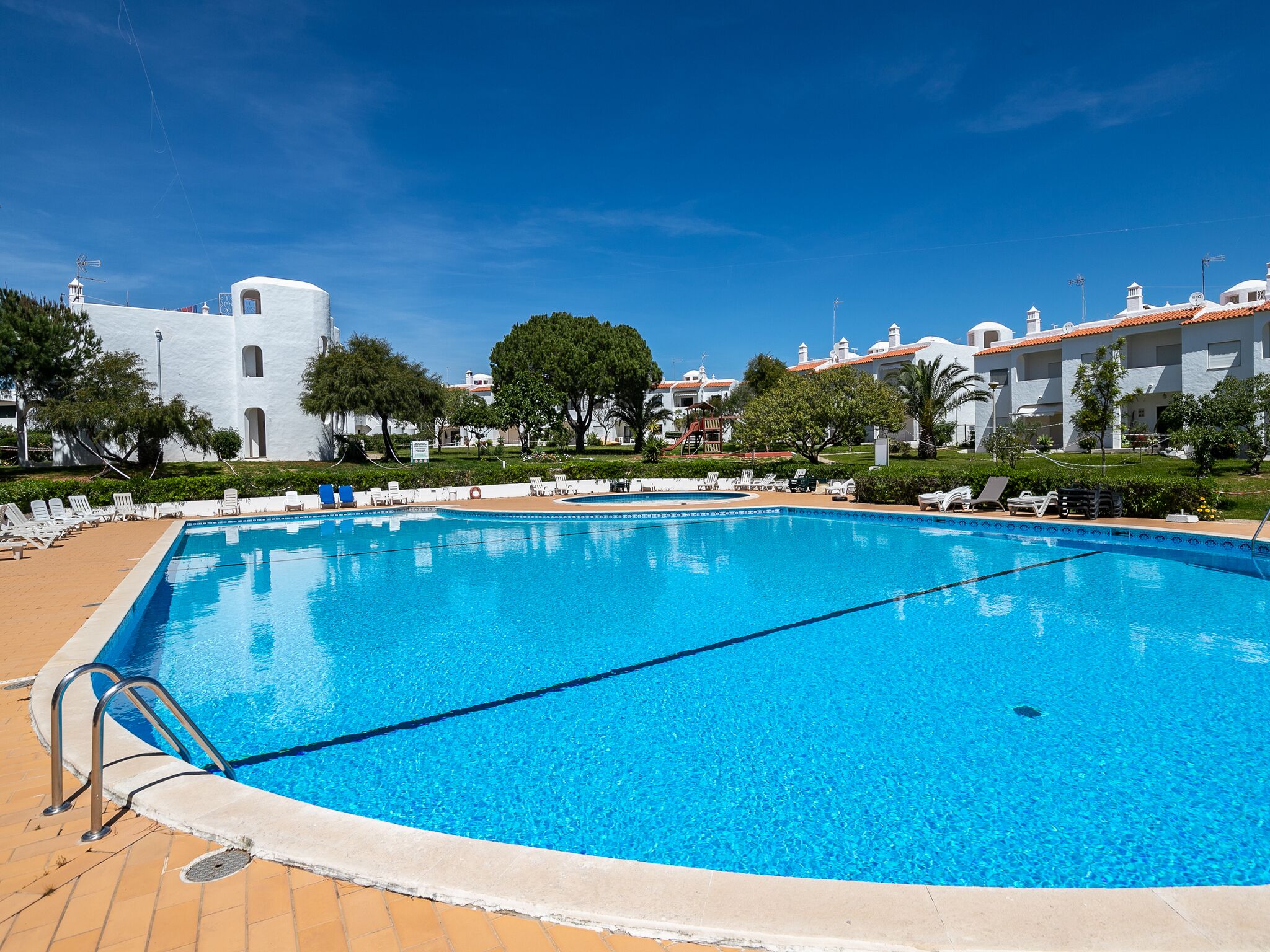 En klarblå pool med en kakelbeklädd poolkant, omgiven av solstolar och en vit byggnad med röda tegeltak i bakgrunden.