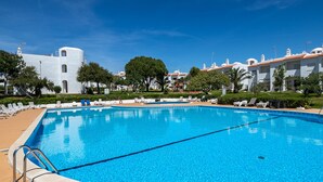Une piscine d’un bleu éclatant, bordée d’une terrasse carrelée, entourée de chaises longues et, au fond, un bâtiment blanc aux toits recouverts de tuiles rouges.