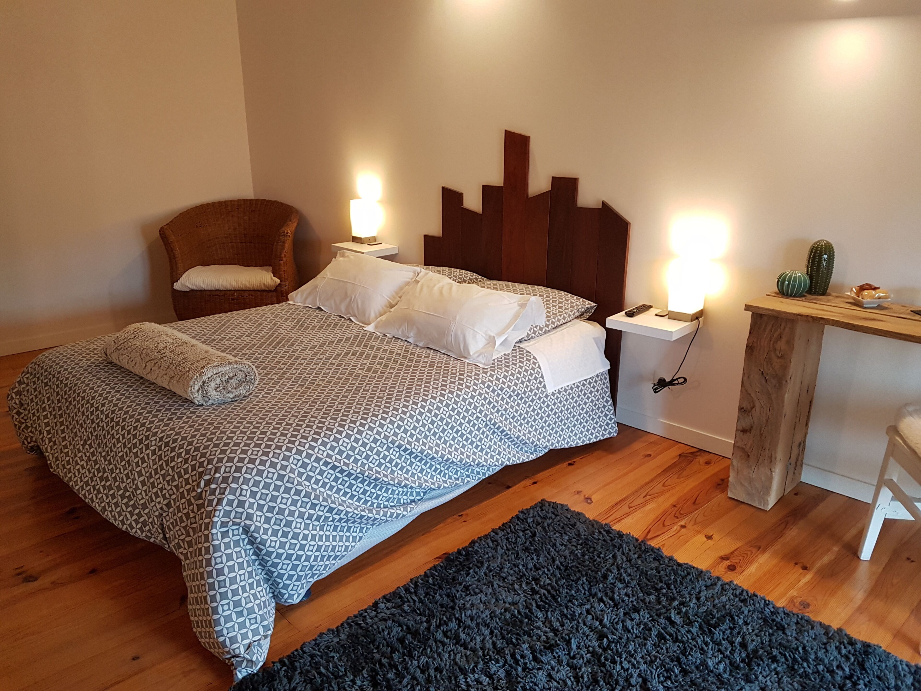A neatly made bed with a patterned bedspread, two pillows, and a rolled towel. There is a wooden nightstand with a lamp, a small cactus, and a decorative item on top. A wicker chair is positioned next to the bed.