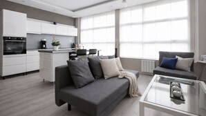 Ein modernes Wohnzimmer mit grauem Sofa, einem Couchtisch und einer Küchenzeile mit weißen Schränken und einem eingebauten Backofen.