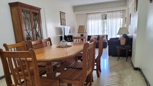 Uma sala de jantar com mesa e cadeiras de madeira, televisão e um sofá ao fundo.