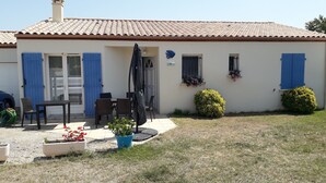 Uma casa com telhado em telhas, um pátio com mesa e cadeiras, uma porta azul e um pequeno jardim com arbustos.