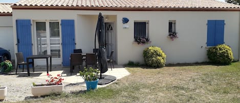 A house with a tiled roof, a patio with a table and chairs, a blue door, and a small garden with bushes.
