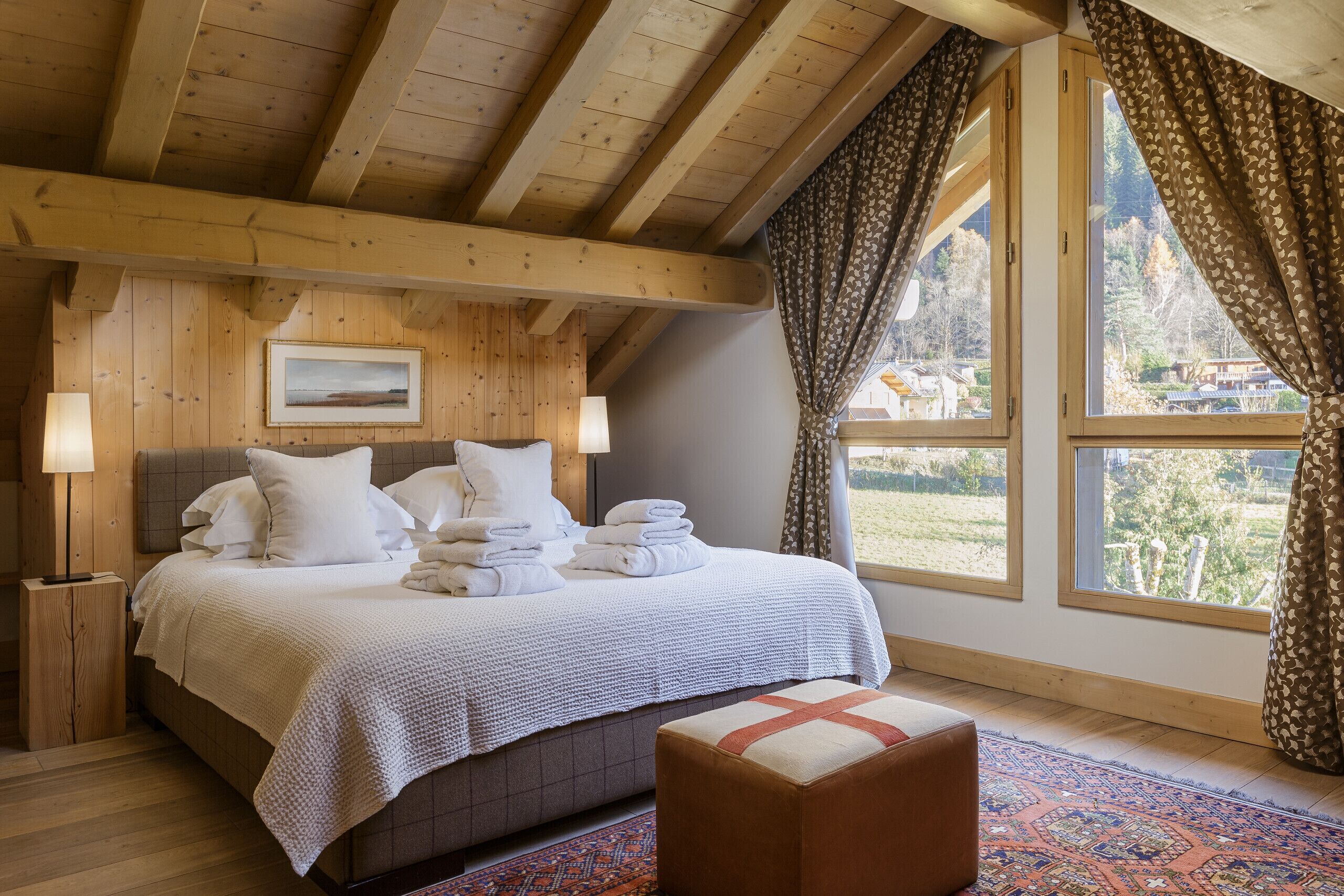 Une chambre avec un plafond en bois, une grande fenêtre avec des rideaux, un lit avec du linge de lit blanc et une table de chevet avec une lampe.