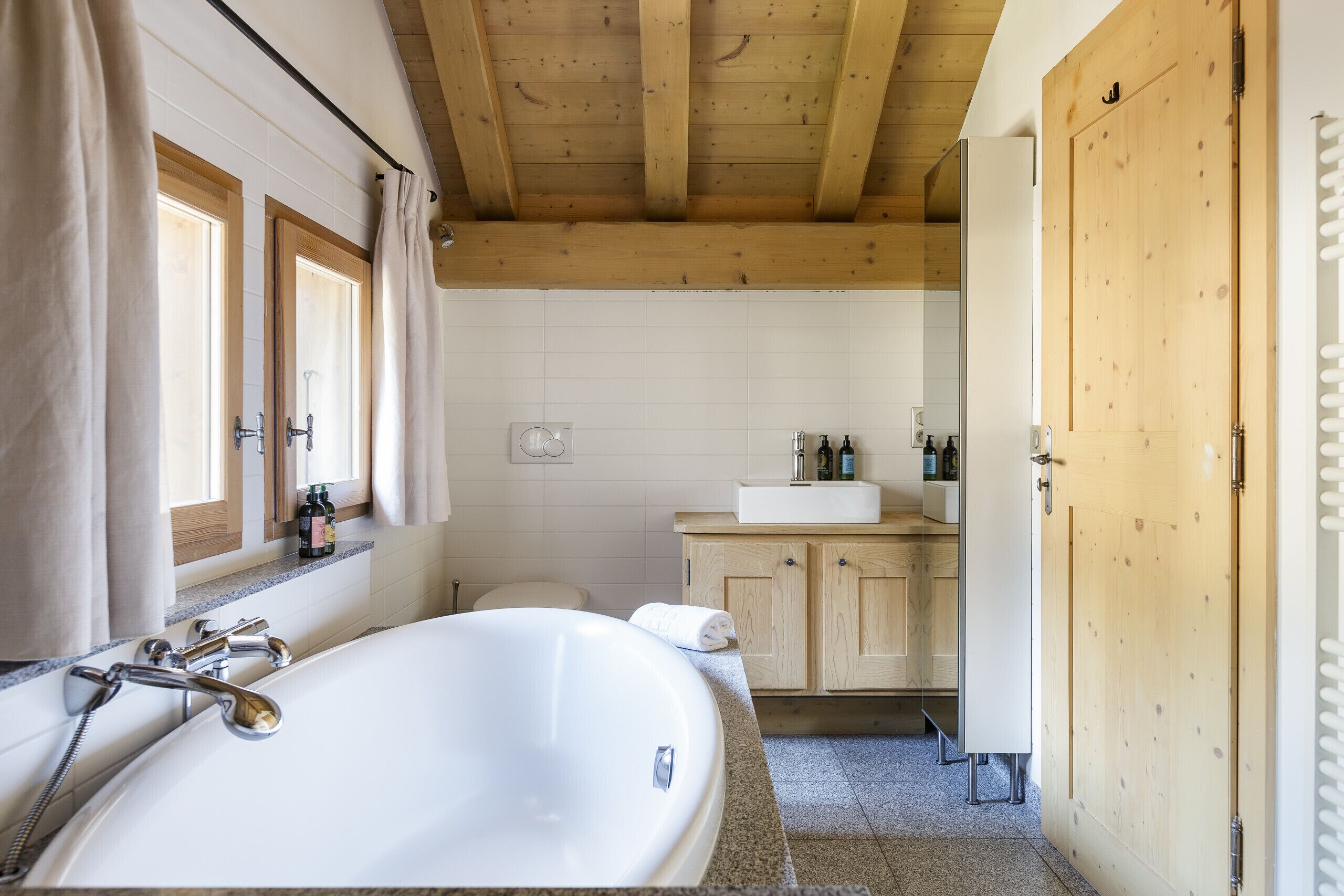 Une salle de bain avec une baignoire sur pieds, des meubles en bois, un lavabo avec des articles de toilette et une fenêtre avec des rideaux.