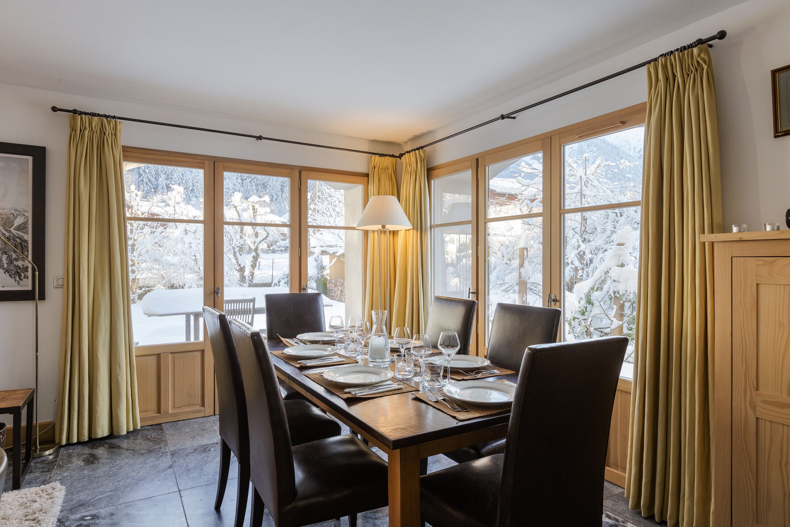 Un coin repas avec une table en bois dressée pour quatre personnes, de grandes fenêtres avec des rideaux et une vue sur des arbres couverts de neige.