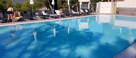 A swimming pool with lounge chairs and umbrellas, surrounded by trees and buildings.