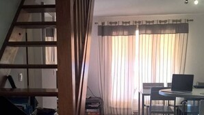 Una escalera conduce a una sala de estar con una mesa de comedor y sillas, una computadora portátil sobre la mesa y una ventana con cortinas.