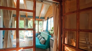 Ein Zimmer mit Holzboden, einem petrolfarbenen Sofa mit Kissen, einem Holzschrank und Blick ins Freie durch ein Fenster mit Vorhängen.
