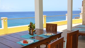 A wooden table set with plates, cutlery, and a bottle of olive oil, overlooking a clear blue sea. - Pretty villa on the island of Maio Cape Verde with panoramic views of  Atlantic (Vila do Maio)