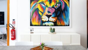 Un comedor con una mesa blanca, sillas, un extintor, una decoración de cactus y un cuadro de un león colorido en la pared.