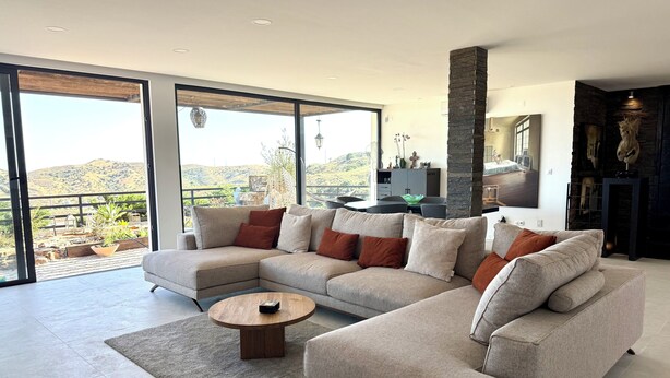 A modern living room with a sectional sofa, a round coffee table, and large sliding glass doors leading to a balcony with a view of hills.