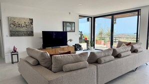 A modern living room with a large flat-screen TV, a grey sofa, and a view of the outdoors through large windows.