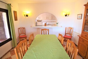 Ein Essbereich mit einem langen Tisch, Holzstühlen, einer durch einen Bogen sichtbaren Küche und Wandleuchten.