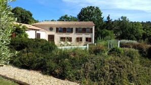 A two-story house with a tiled roof, surrounded by dense greenery and a gravel path.