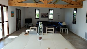 A spacious living room with a high ceiling, a large window, a dark leather sofa, a white dining table, and a TV on a stand.