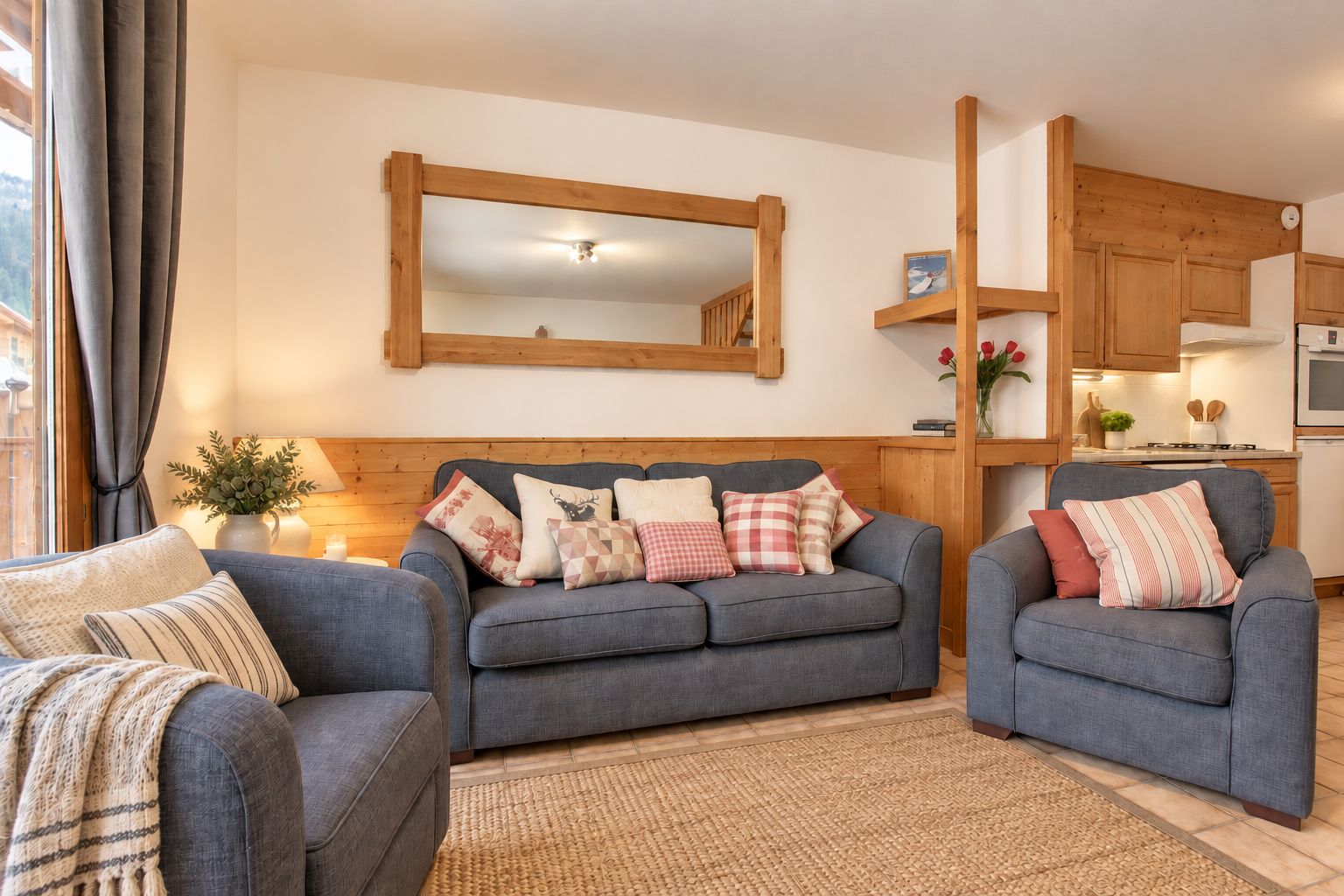 A living room with a grey sofa set, a wooden-framed mirror, and a kitchen area with wooden cabinets and a stove.