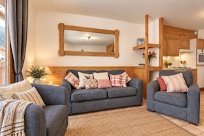 A living room with a grey sofa set, a wooden-framed mirror, and a kitchen area with wooden cabinets and a stove. - Telemark Two. Perfect location in the centre of town. (Morzine)