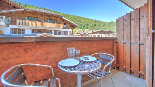 Varanda de madeira com mesa e cadeiras, com vista para uma área montanhosa e edifícios tradicionais.