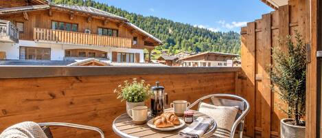 Un balcon en bois avec une table préparée pour le petit-déjeuner, comprenant des croissants, du café et une plante.