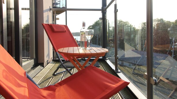 Un balcone con ringhiera in vetro, una sedia rossa, un piccolo tavolo di legno e una bottiglia di vino.