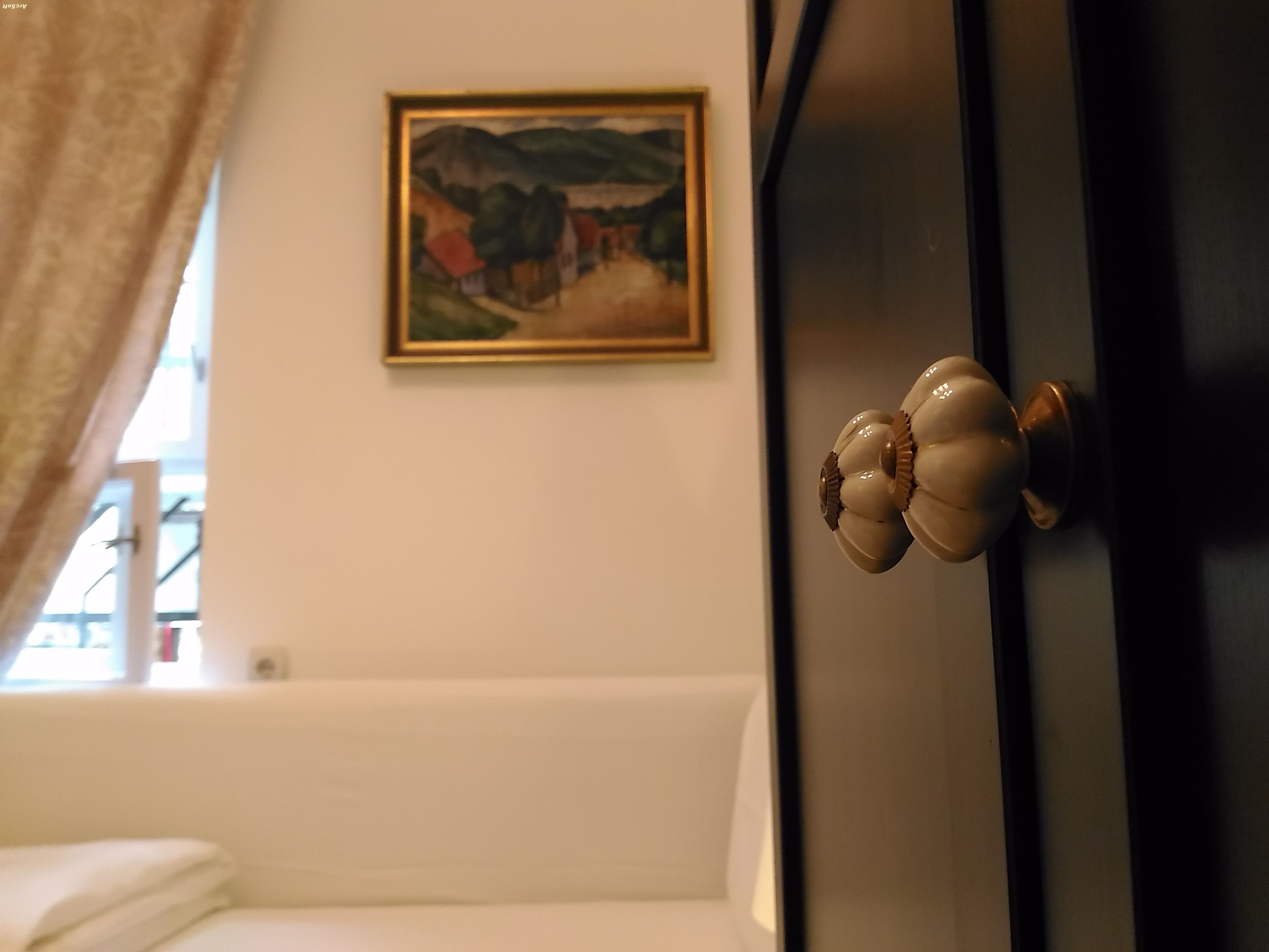 A room with a bed, a door with a decorative knob, a framed painting on the wall, and a window with curtains.