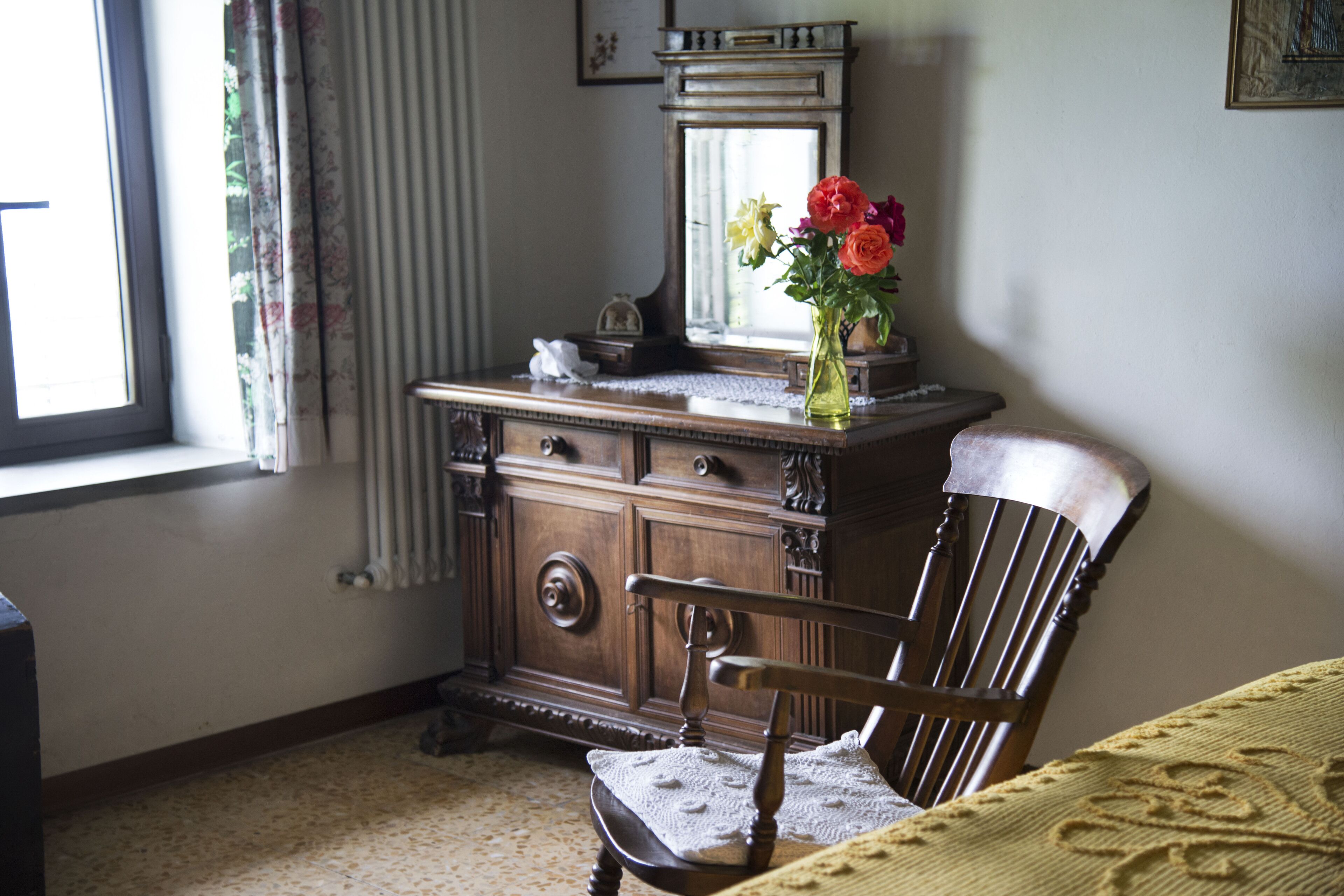 Un aparador de madera con un espejo, una silla con mantel de encaje y un florero con flores.