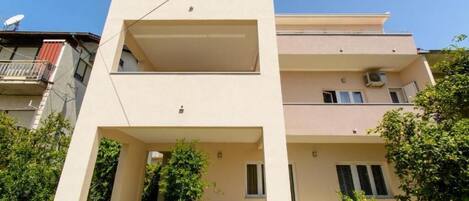 A modern two-story building with a flat roof, a balcony on the upper floor, and a small garden area in front.