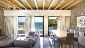 A modern living room with a sofa, dining table, and chairs. There is a view of the ocean through large windows. - Villa Passithea , 3 bedroom villa plus studio with private heated pool (Lindos)