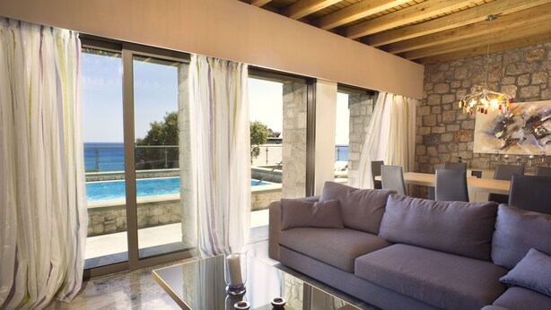 A modern living room with a large sofa, a glass coffee table, and a view of the ocean through large windows. - Villa Passithea , 3 bedroom villa plus studio with private heated pool (Lindos)