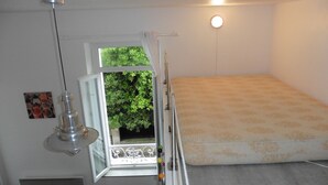 Een kleine kamer met een enkel bed, een raam met uitzicht op groen, een hanglamp en een klein prentje aan de muur.