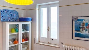 A room with a white cabinet containing colorful mugs and cups, a window with a white frame, and a painting of a lighthouse on the wall. - Superior Apartment Sea View (Polignano A Mare)