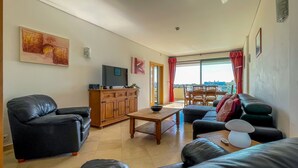 A living room with a TV, wooden cabinet, leather sofas, and a dining area with a view.
