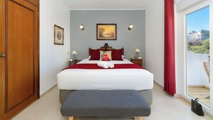 A bedroom with a bed, a bench, a wardrobe, a painting, and a view through a window. - (^ ‿ ^) Cozy & Bright, near the Beach, Marina, Historic Center and Supermarket (Lagos)