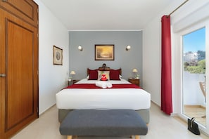 A bedroom with a bed, a bench, a wardrobe, a painting, and a view through a window.