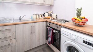 A modern kitchen with light wood cabinets, a stainless steel sink, a built-in oven, a washing machine, and a fruit bowl on the countertop. - One Bedroom Apartment by Klass Living Serviced Accommodation Rutherglen - Crossroads Apartment With WiFi and Parking (Glasgow)