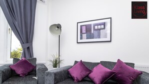 A living room with two grey sofas, each with a purple cushion, a floor lamp, a framed picture on the wall, and a window with curtains. - One Bedroom Apartment by Klass Living Serviced Accommodation Rutherglen - Crossroads Apartment With WiFi and Parking (Glasgow)