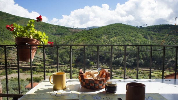 A balcony with a table set for breakfast, including a basket of bread, a mug, and a flower-patterned placemat.