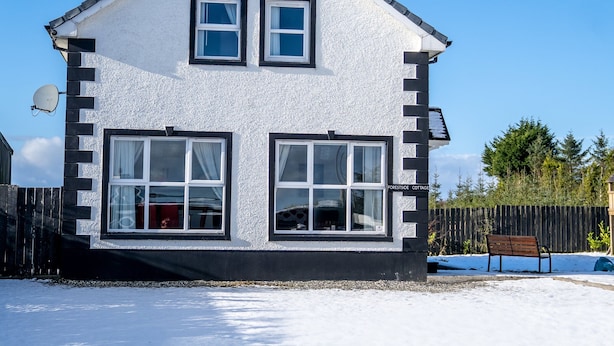 A two-story cottage with a white exterior, two windows on the upper floor, and a large window on the ground floor.