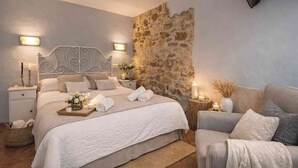Un dormitorio con pared de piedra, cama con ropa de cama blanca, mesita de noche con lámpara y una silla.
