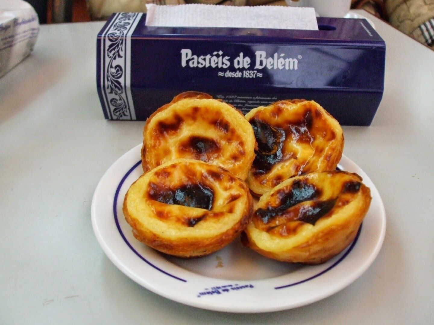 A plate of Pastéis de Belém pastries on a white table with a box of the same name in the background.
