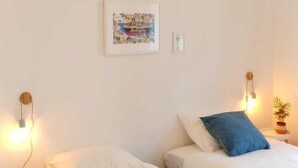 Ett rum med två sängar, en hängande lampa, en tavla som hänger på väggen och ett litet bord med en växt.
