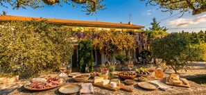 Un área de comedor al aire libre con una variedad de comida y bebidas, rodeada de vegetación y un edificio con tejado de azulejos.