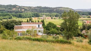 Un paysage rural avec un bâtiment blanc surmonté d’un toit recouvert de tuiles rouges, entouré de verdure et avec des collines en arrière-plan.