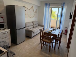 Une cuisine compacte équipée d’un réfrigérateur, d’un four à micro-ondes et d’un lave-vaisselle. On y trouve également un canapé et une table à manger avec des chaises. Un extincteur est visible sur le mur.