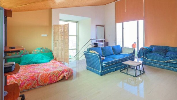 A spacious living room with a wooden ceiling, a sofa set, a bed with a colorful bedspread, and a staircase leading to an upper level.