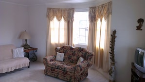 A living room with a floral sofa, a small table with a lamp, a television on a stand, and curtains on the windows.