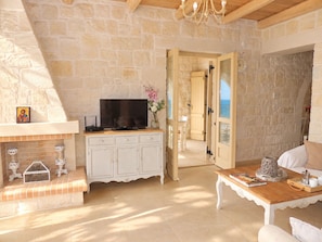 Une pièce aux murs de pierre, avec un téléviseur posé sur un meuble, une table avec des livres et un plateau, et une vue sur l’extérieur par une porte ouverte.