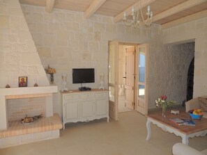 A room with a stone wall, a fireplace, a television, a wooden cabinet, and a table with a bowl of fruit. - A charming 3 bedroom stone villa w/ privacy, private pool & fantastic sea views. (Chania)