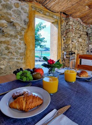 Una mesa de desayuno con croissants, fruta y jugo, frente a una pared de piedra con vista a la vegetación.